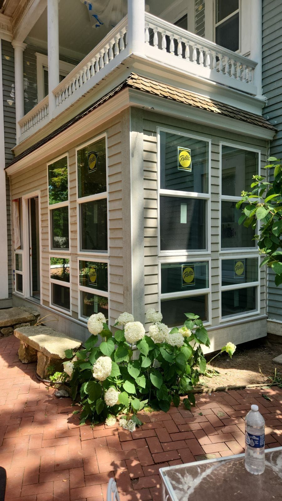 A sunroom addition with light gray siding and many windows, next to a brick patio with a large white hydrangea bush.
