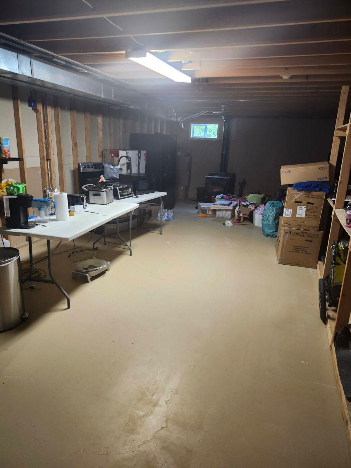 A basement with a long white table, a wood stove, and stacks of cardboard boxes on a concrete floor under fluorescent light.