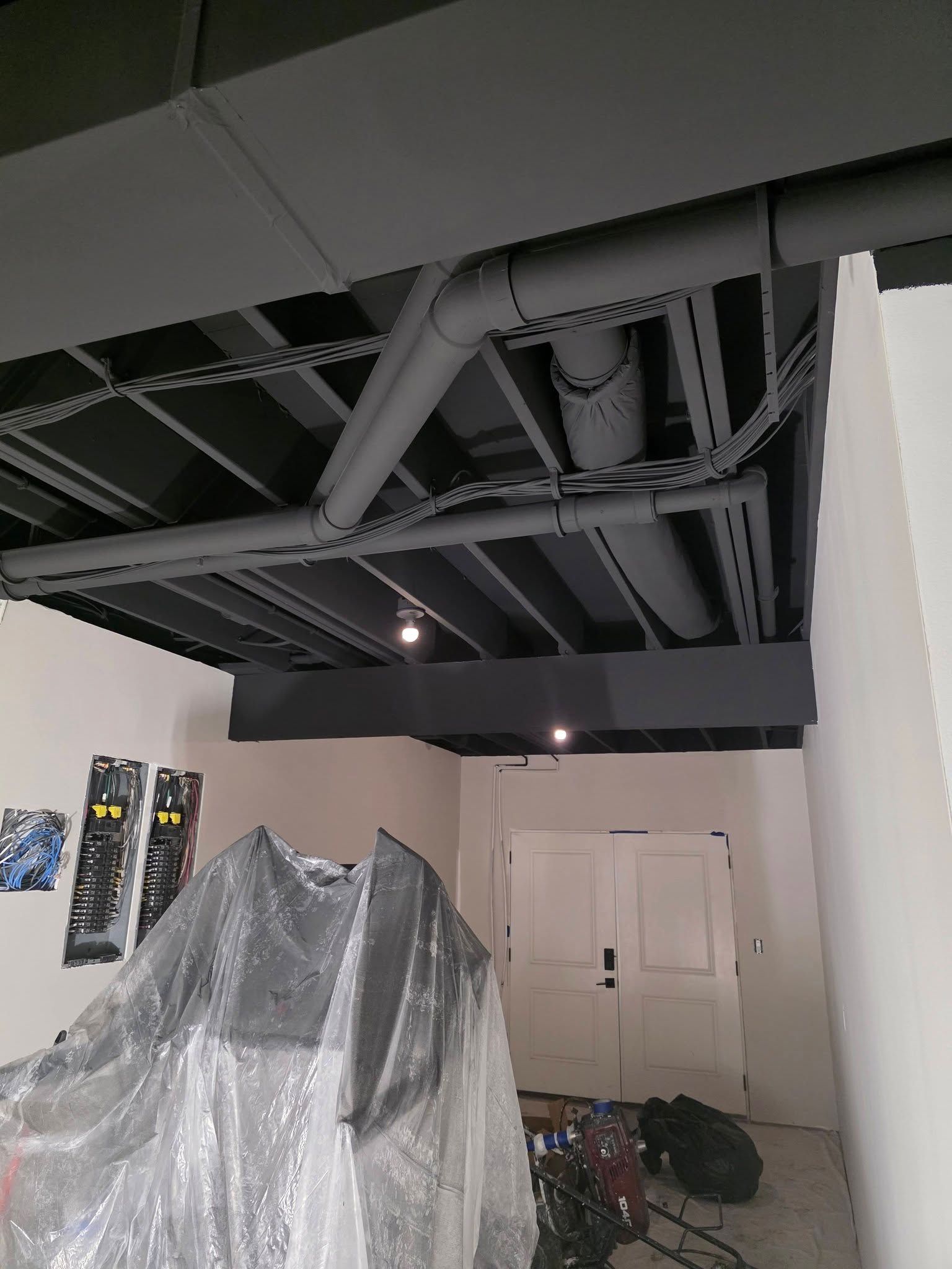 An unfinished basement ceiling painted dark gray with exposed pipes and wiring above a room covered in plastic sheeting.