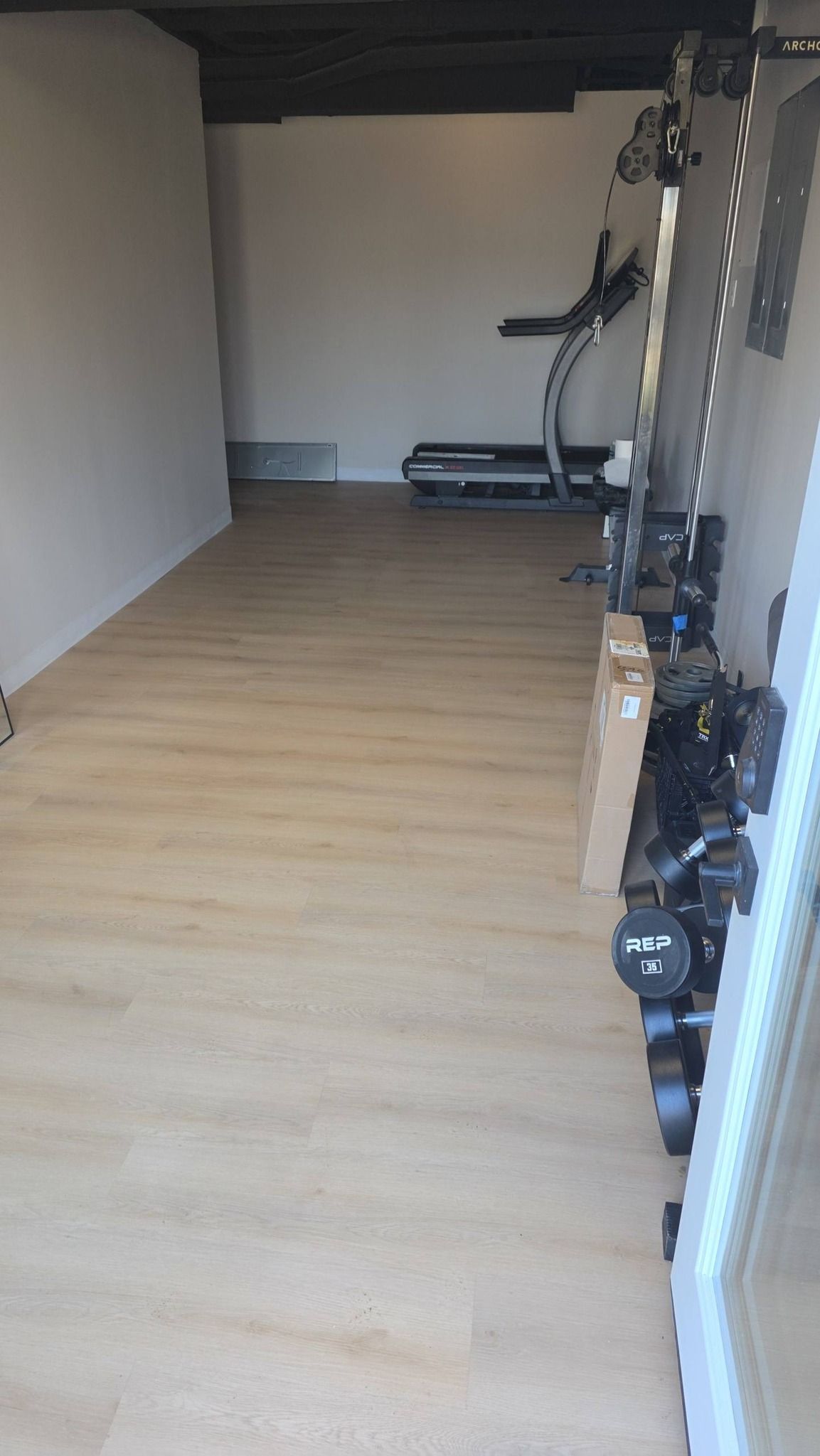 A narrow home gym space with light wood-look flooring, a treadmill in the back, and stacked weights along the right wall.