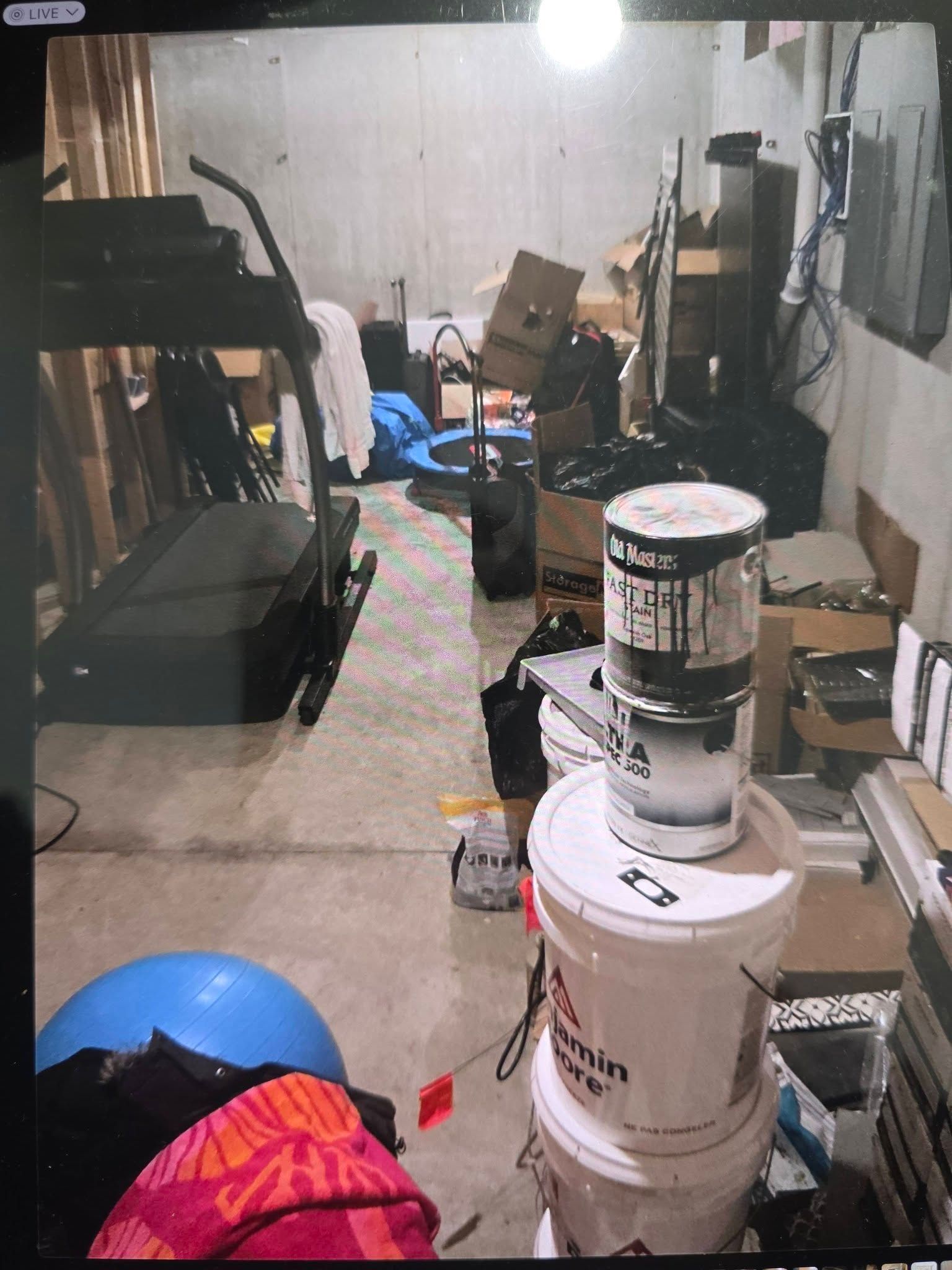 A cluttered basement storage area with a treadmill, stacks of paint buckets, boxes, and miscellaneous items on the floor.