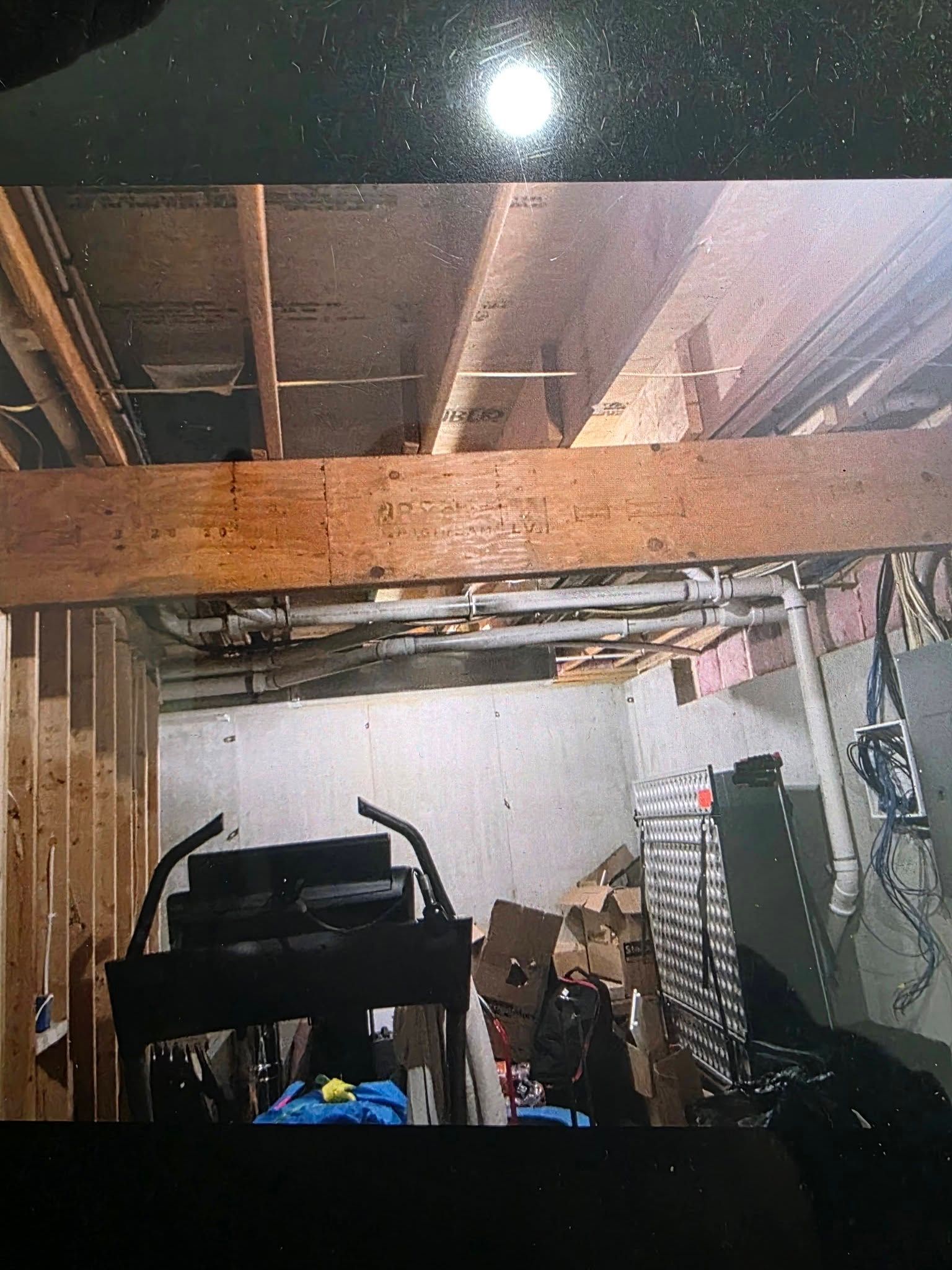 A basement space showing a large wooden support beam, exposed ceiling joists, plumbing pipes, and scattered storage items.