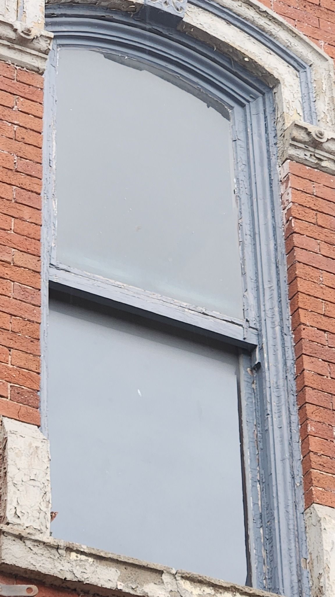 A tall, arched window with a gray-painted wooden frame set into a red brick building facade.