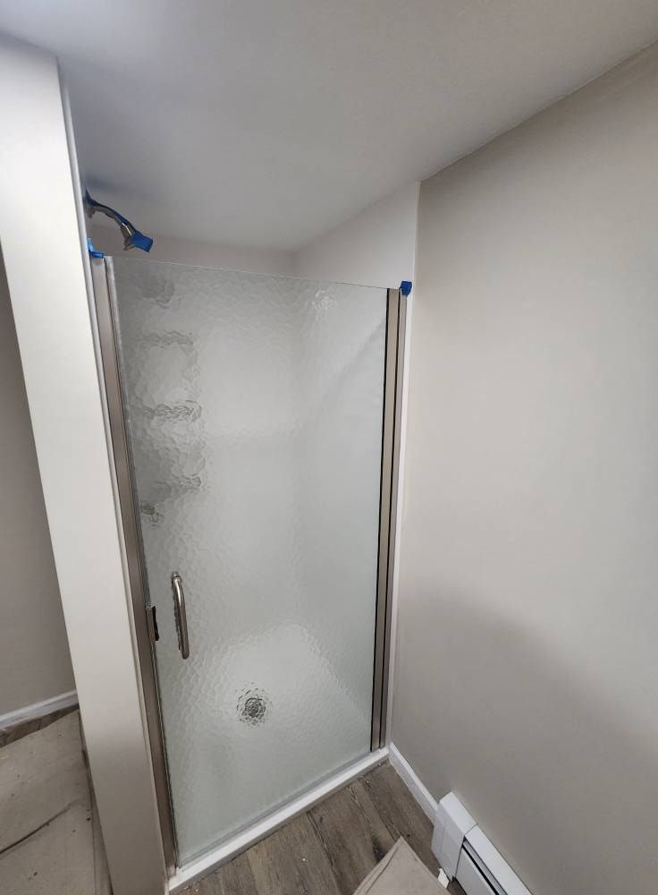 A glass shower door installed between two light-colored walls, featuring a metal frame and handle over wood-look flooring.