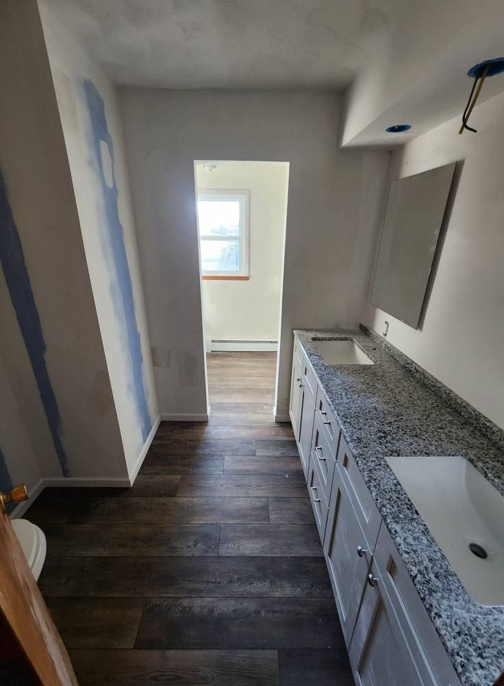 A bathroom under renovation with a double vanity, speckled granite countertop, and wood-look flooring leading to a doorway.