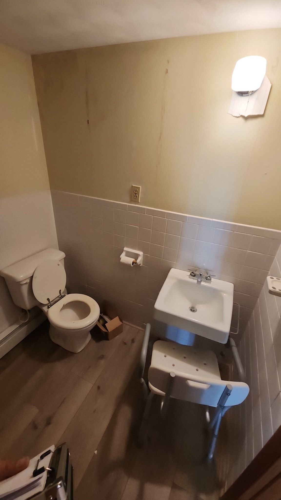 A small bathroom with a toilet and wall-mounted sink, featuring a shower chair placed directly beneath the sink.