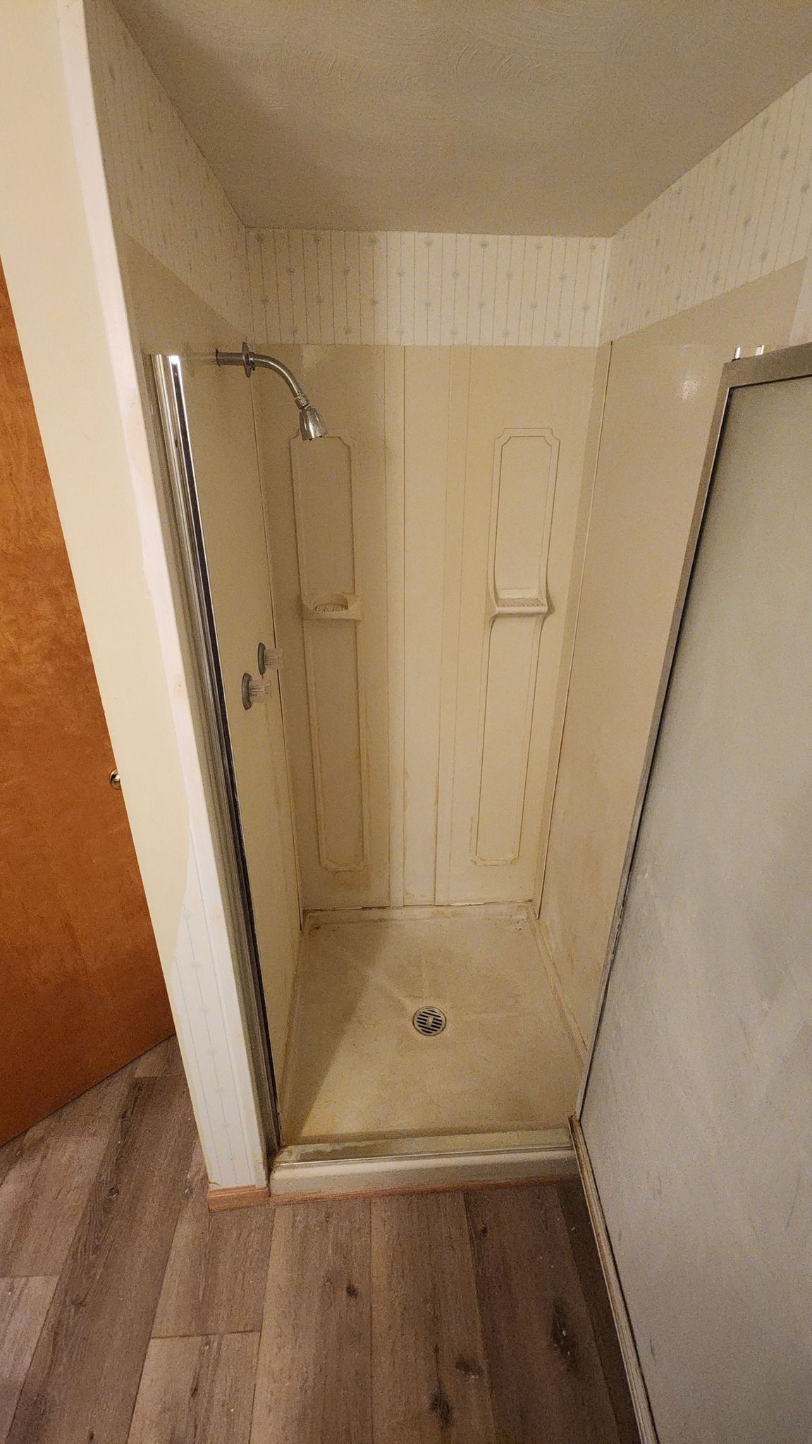 A beige, stall-style shower with an overhead showerhead and a light-colored tiled floor, viewed from a room with flooring.