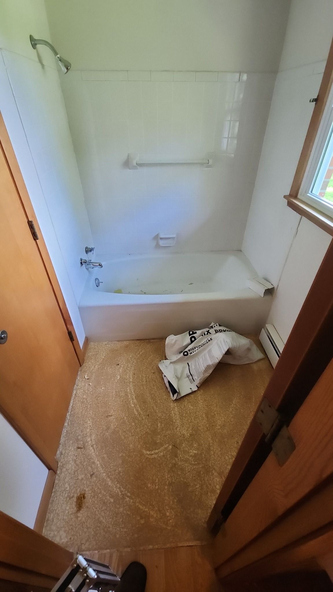 A small, empty bathroom featuring a white bathtub, tiled shower walls, wood-toned door frames, and a floor undergoing repair.