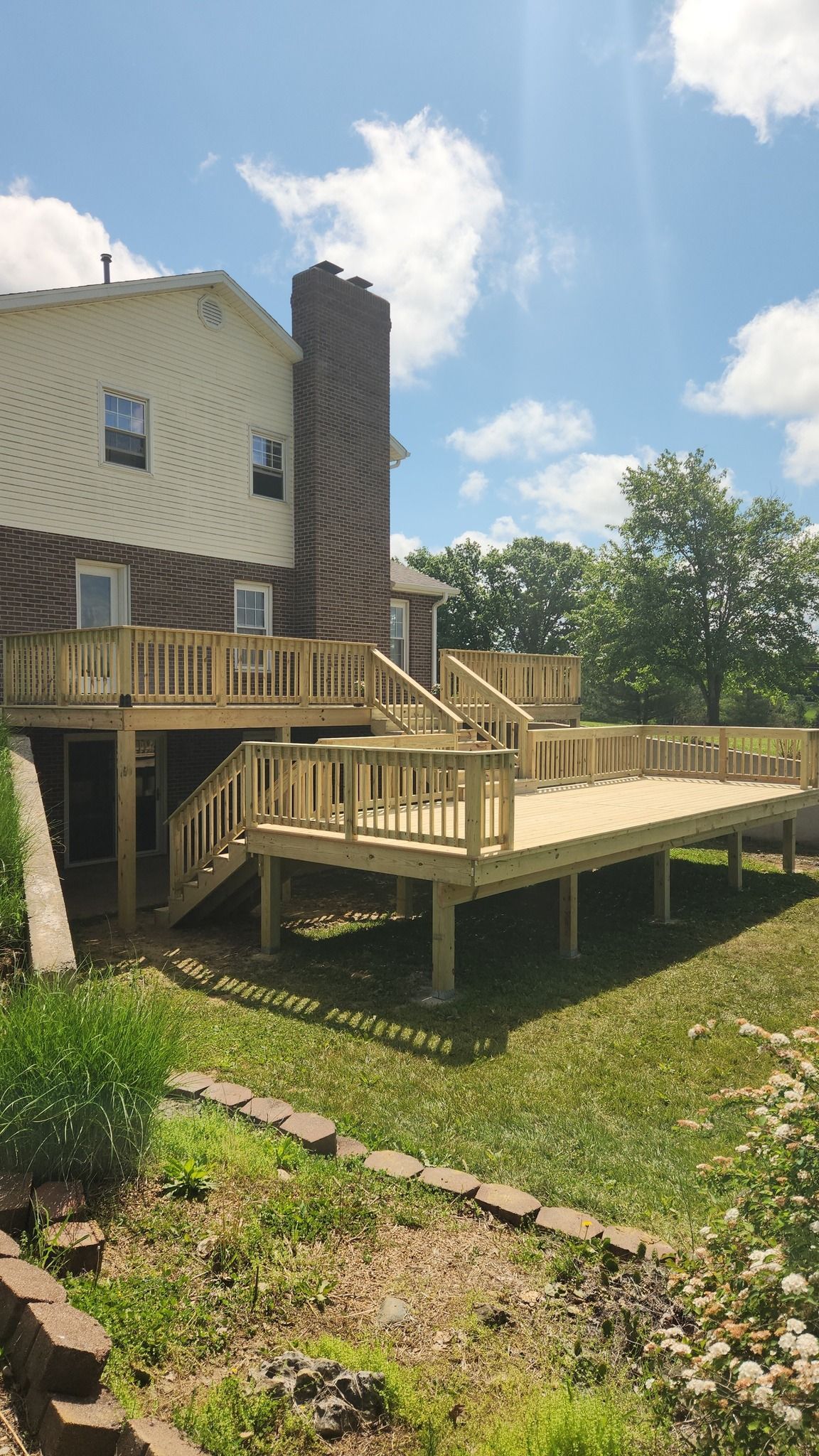 A multi-level light wood deck attached to the back of a brick and light-sided house under a blue sky with scattered clouds.