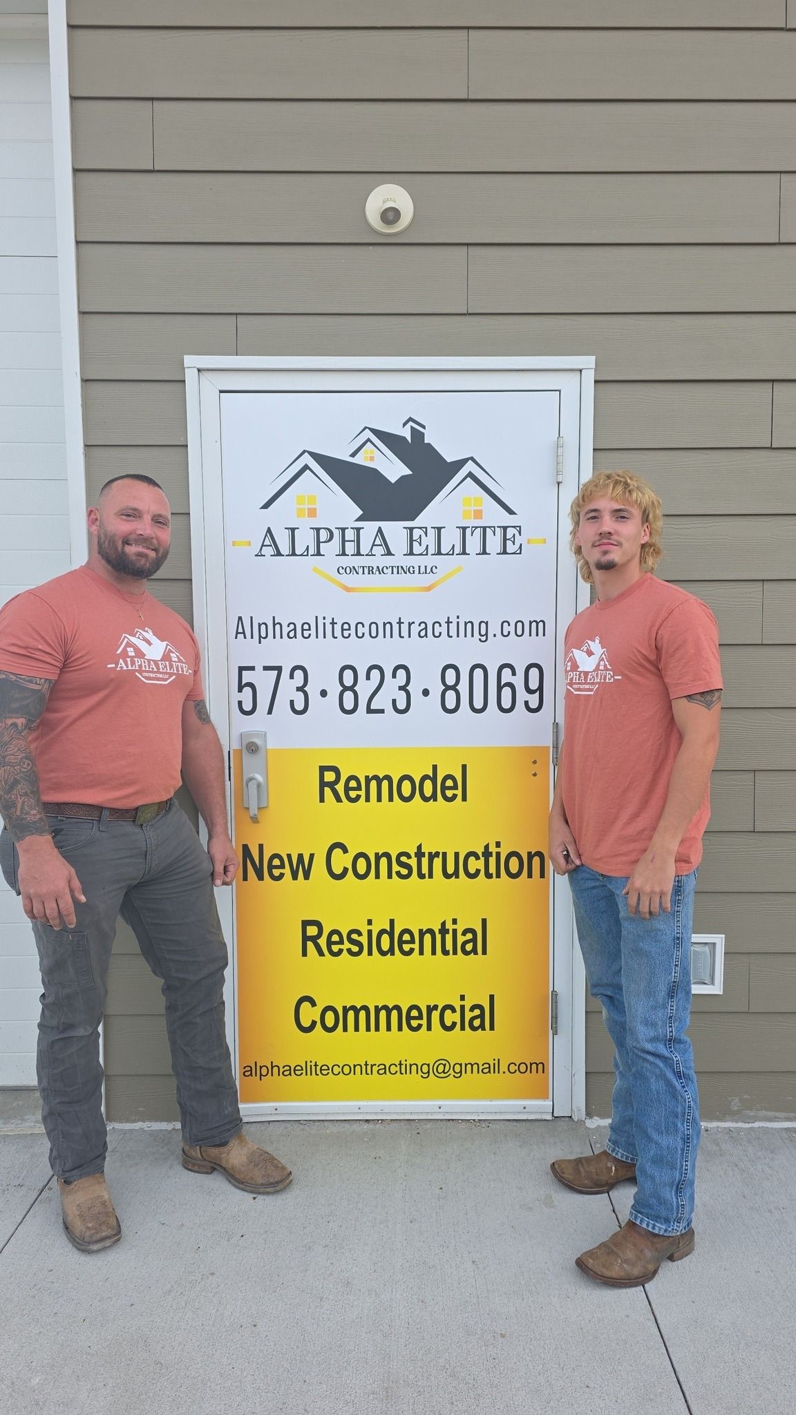 Two construction workers in matching orange shirts stand outside a door labeled Alpha Elite with services and phone number.