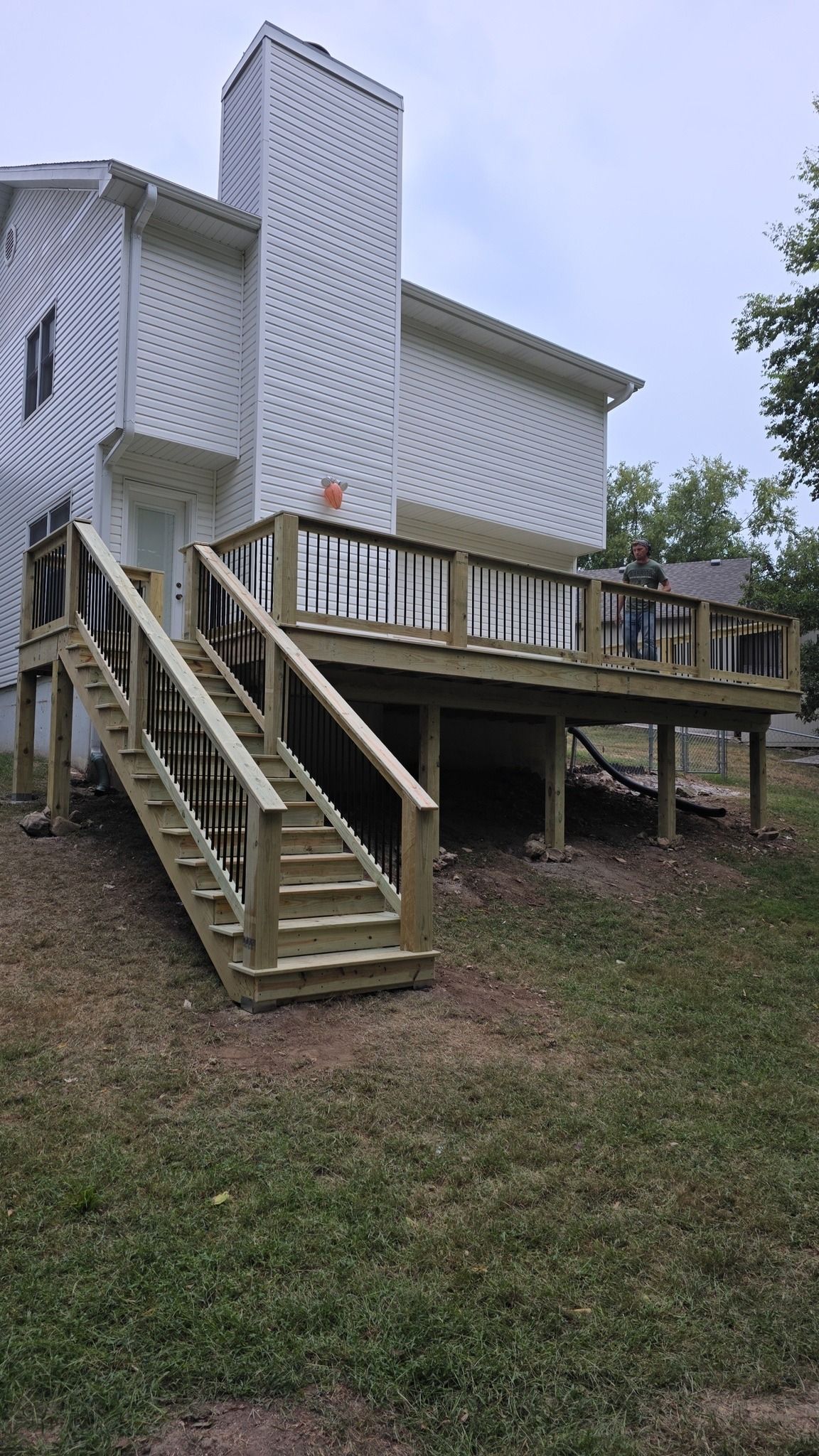 A newly constructed wooden deck and staircase attached to the back of a two-story white house on a grassy lawn.