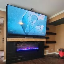 A large TV mounted above a modern electric fireplace with purple flames, set against a dark wall in a living room.