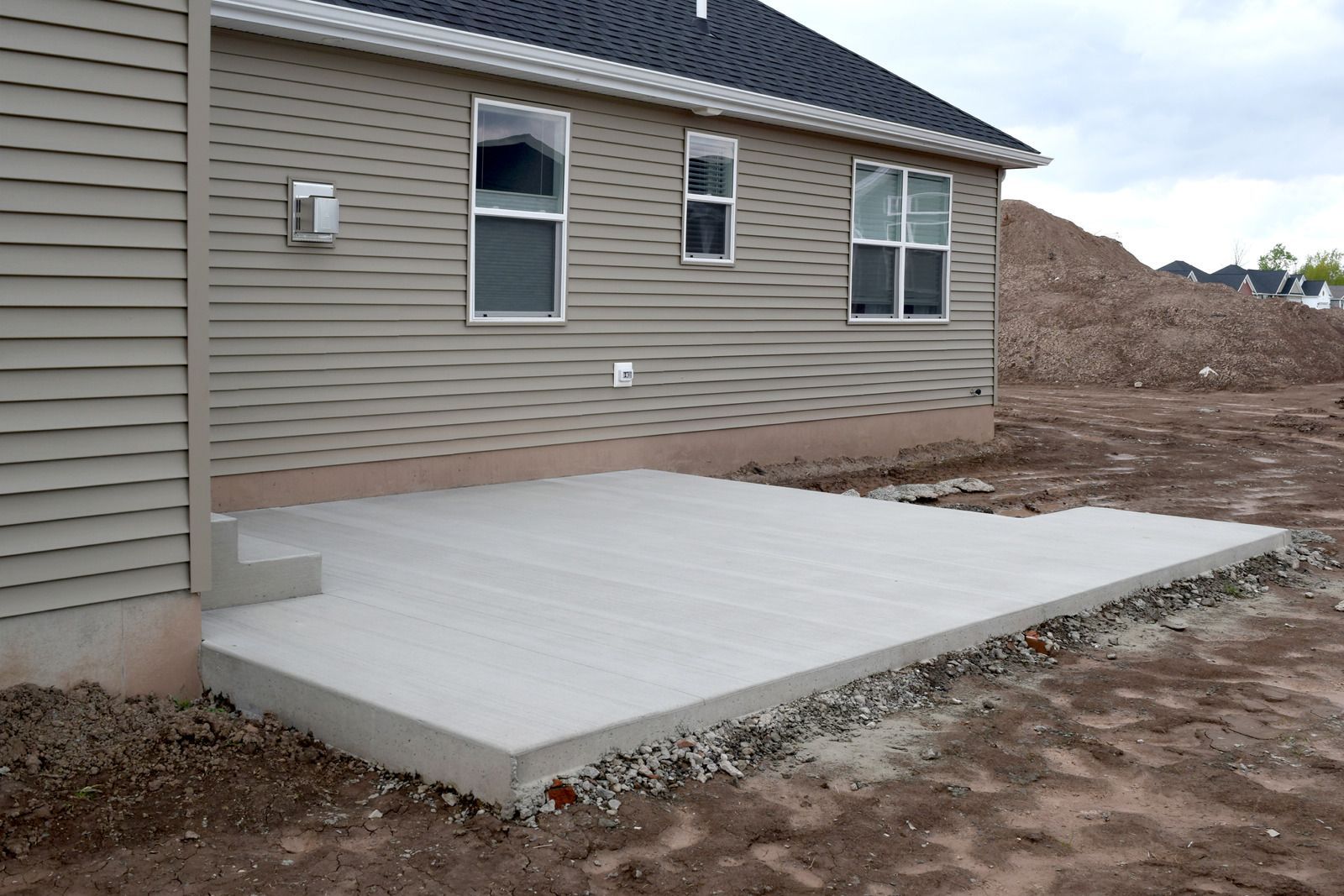 A newly poured concrete patio attached to the side of a beige vinyl-sided house with gravel exposed along the edge.