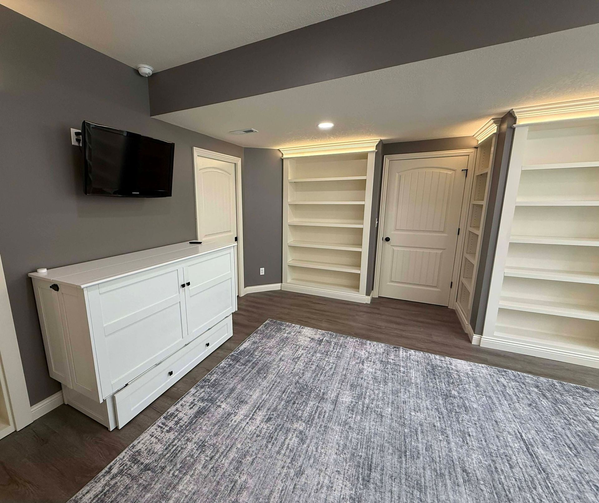 A room with gray walls, a white dresser topped by a wall-mounted TV, built-in white shelves, and a gray patterned rug.