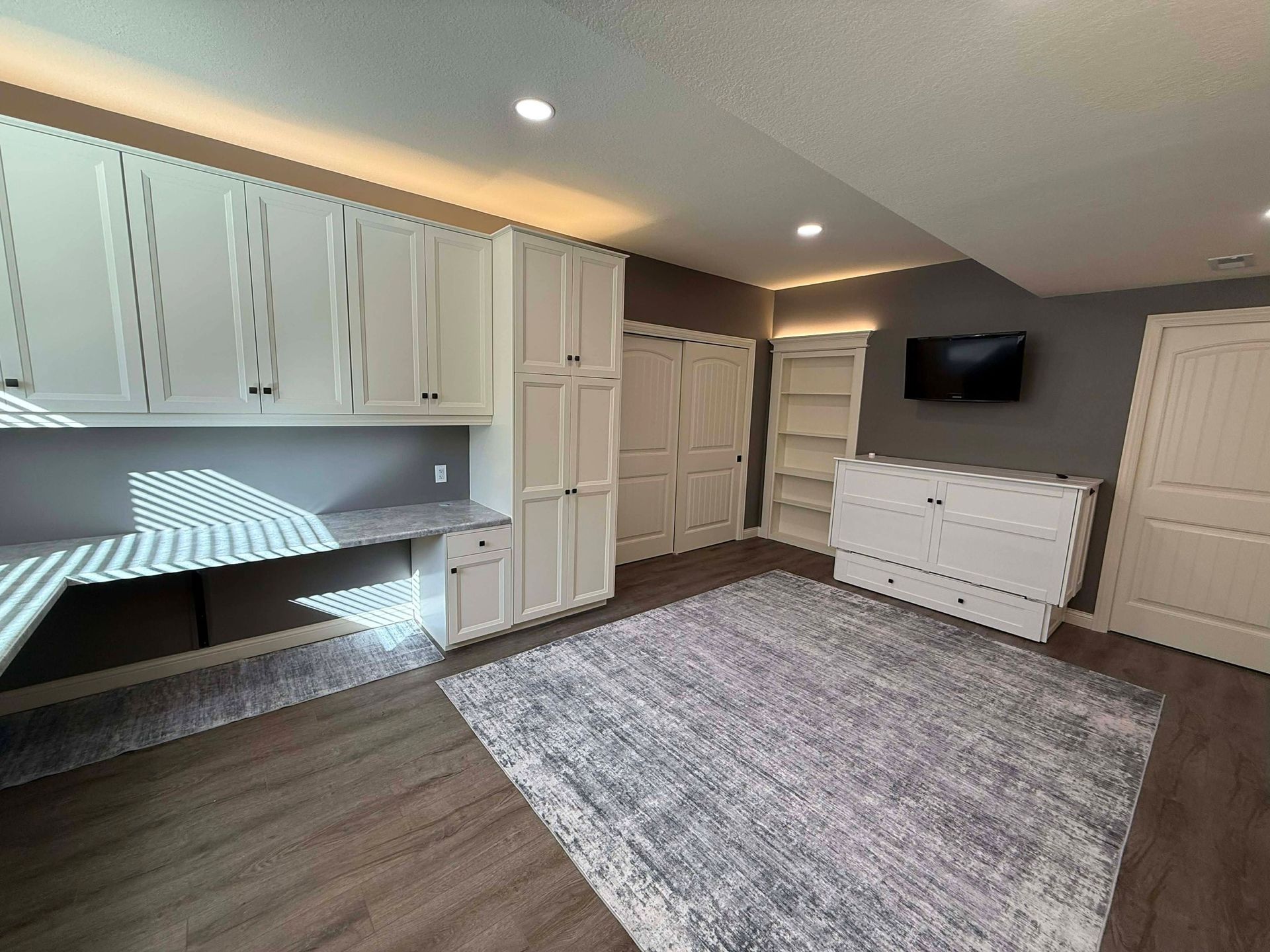 An office space with white cabinets, a desk, a grey patterned rug on wood flooring, and a mounted TV on a dark wall.