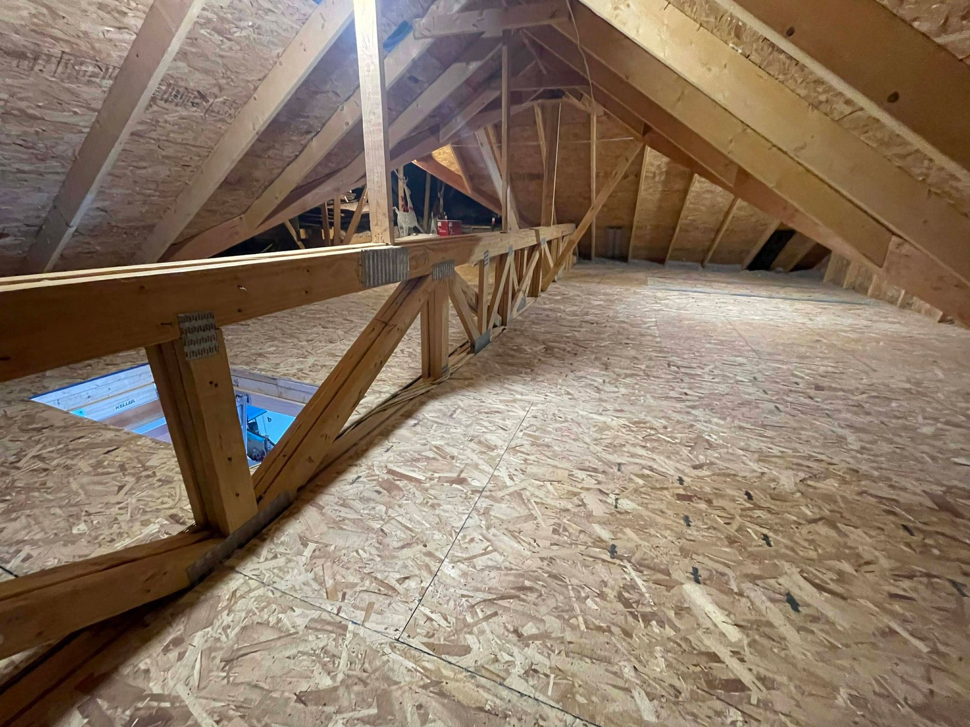 An unfinished attic with wooden roof trusses and an oriented strand board (OSB) floor, showing a small roof hatch.