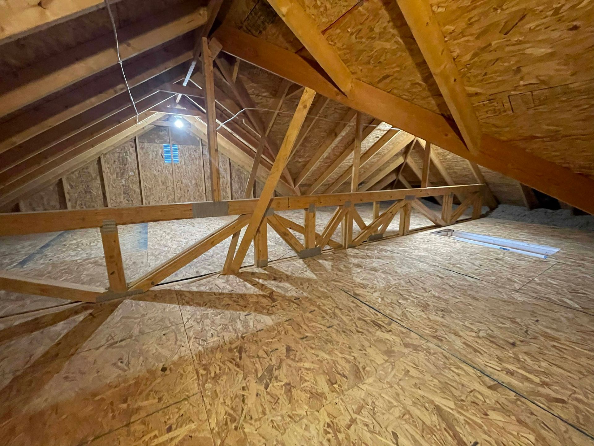 An attic space featuring wooden trusses, OSB flooring, and visible roof framing under ambient lighting.