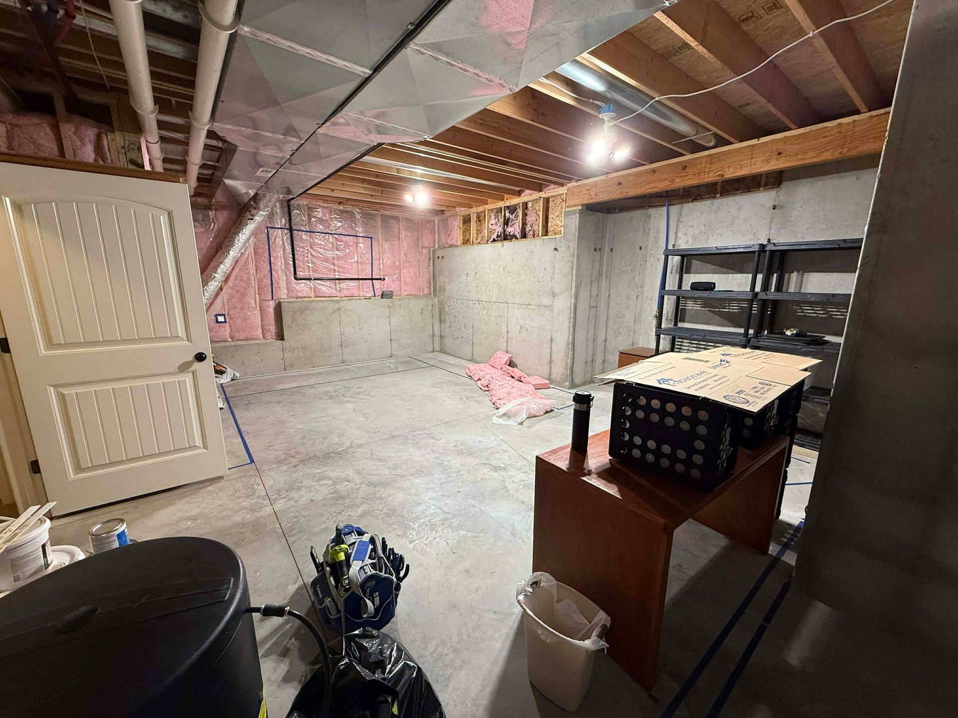 An unfinished basement with concrete walls, exposed ceiling joists, a white door, shelving, and a desk with storage bins.