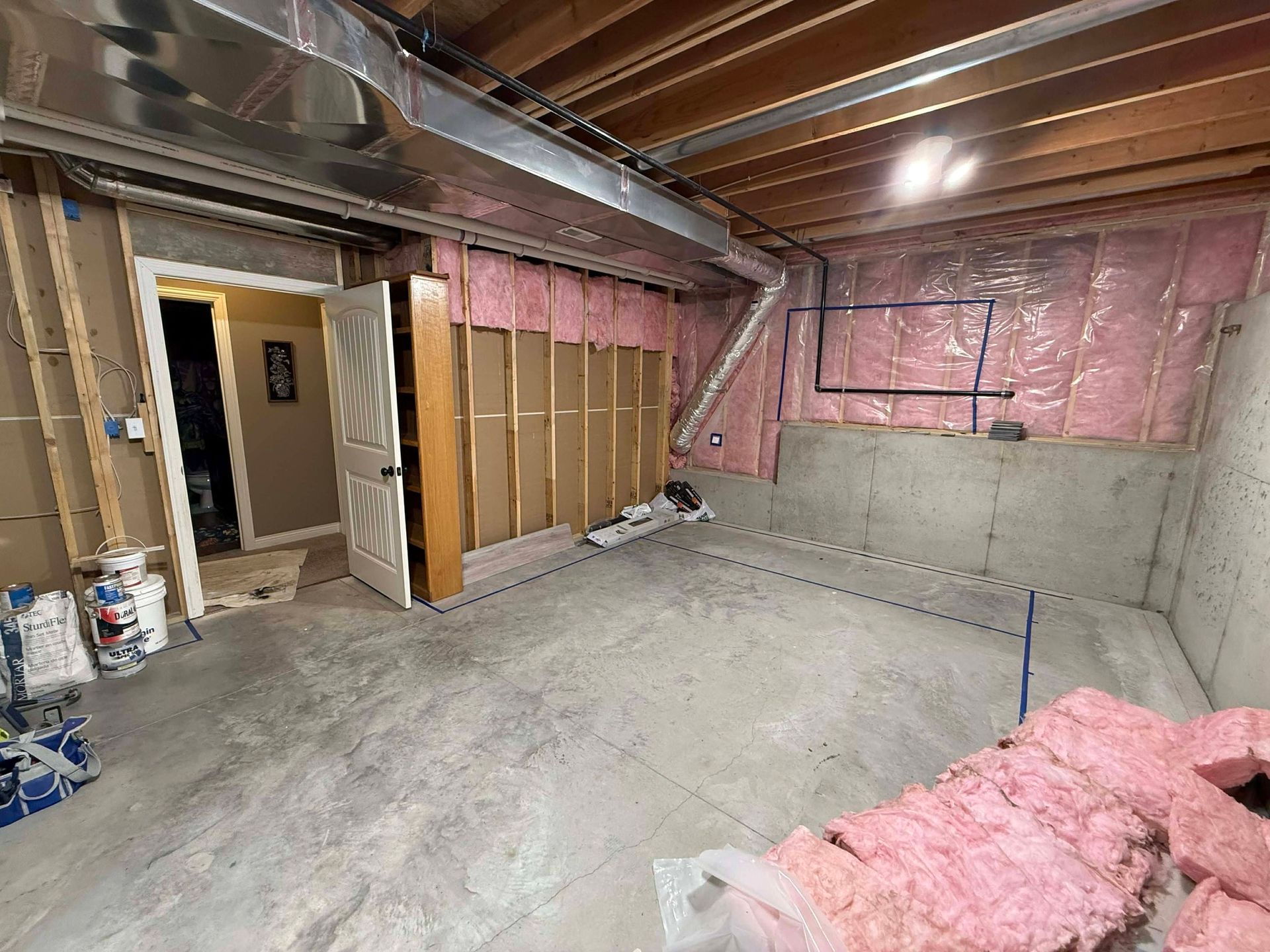 An unfinished basement with exposed wooden studs, pink wall insulation, a concrete floor, and an open white doorway.