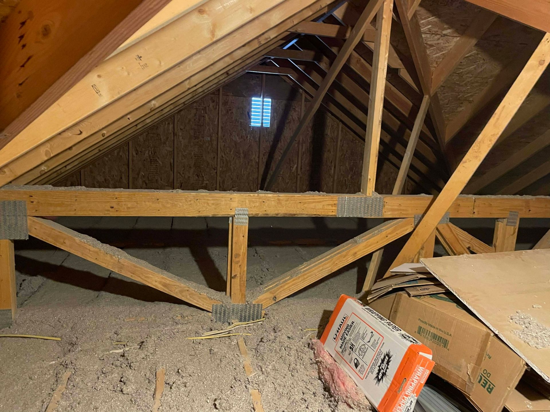An attic interior featuring wooden roof trusses, insulation on the floor, and a small vent on the distant plywood wall.