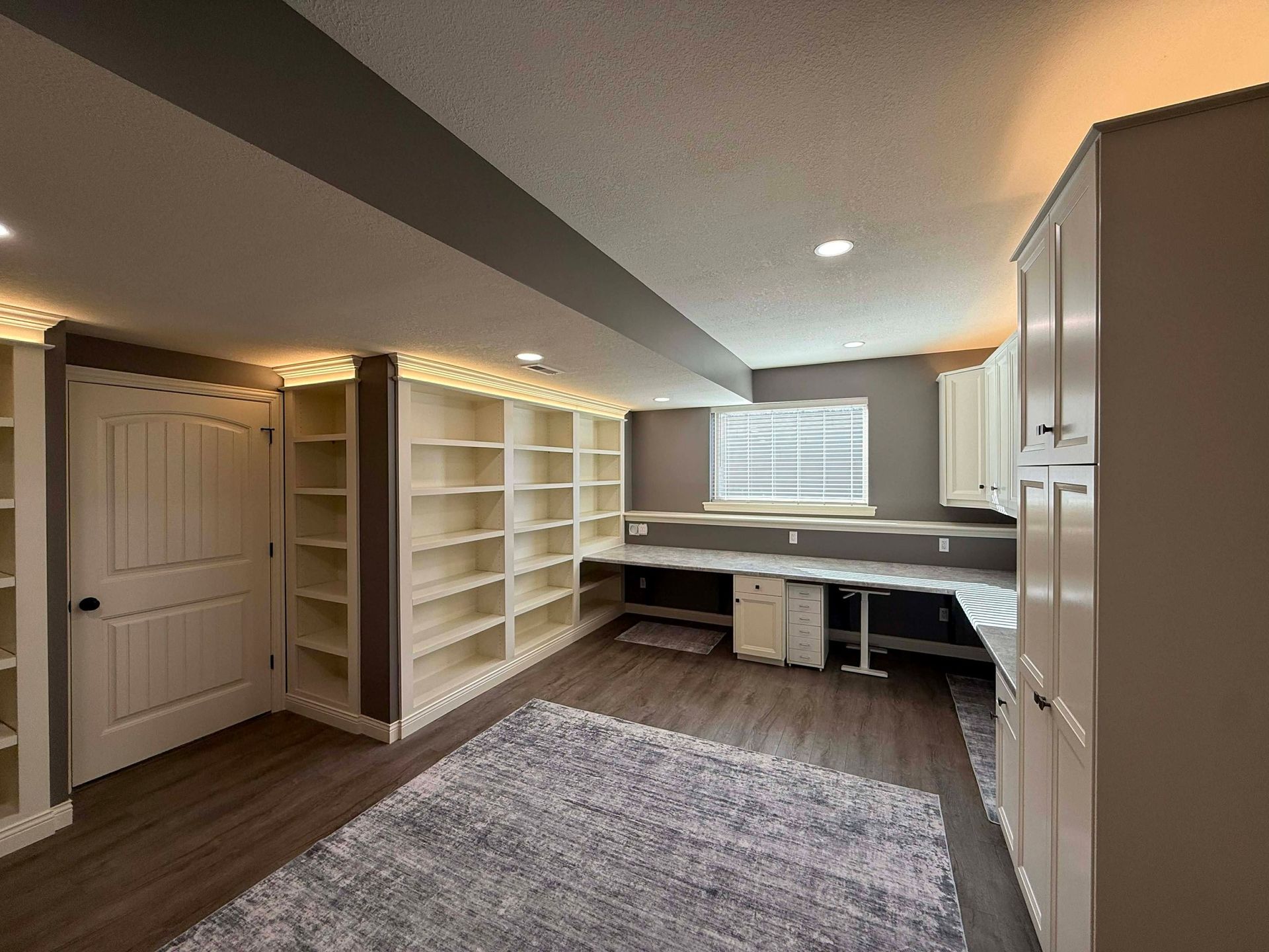 A home office with white built-in shelves, a corner desk, wood flooring, and a large patterned area rug.