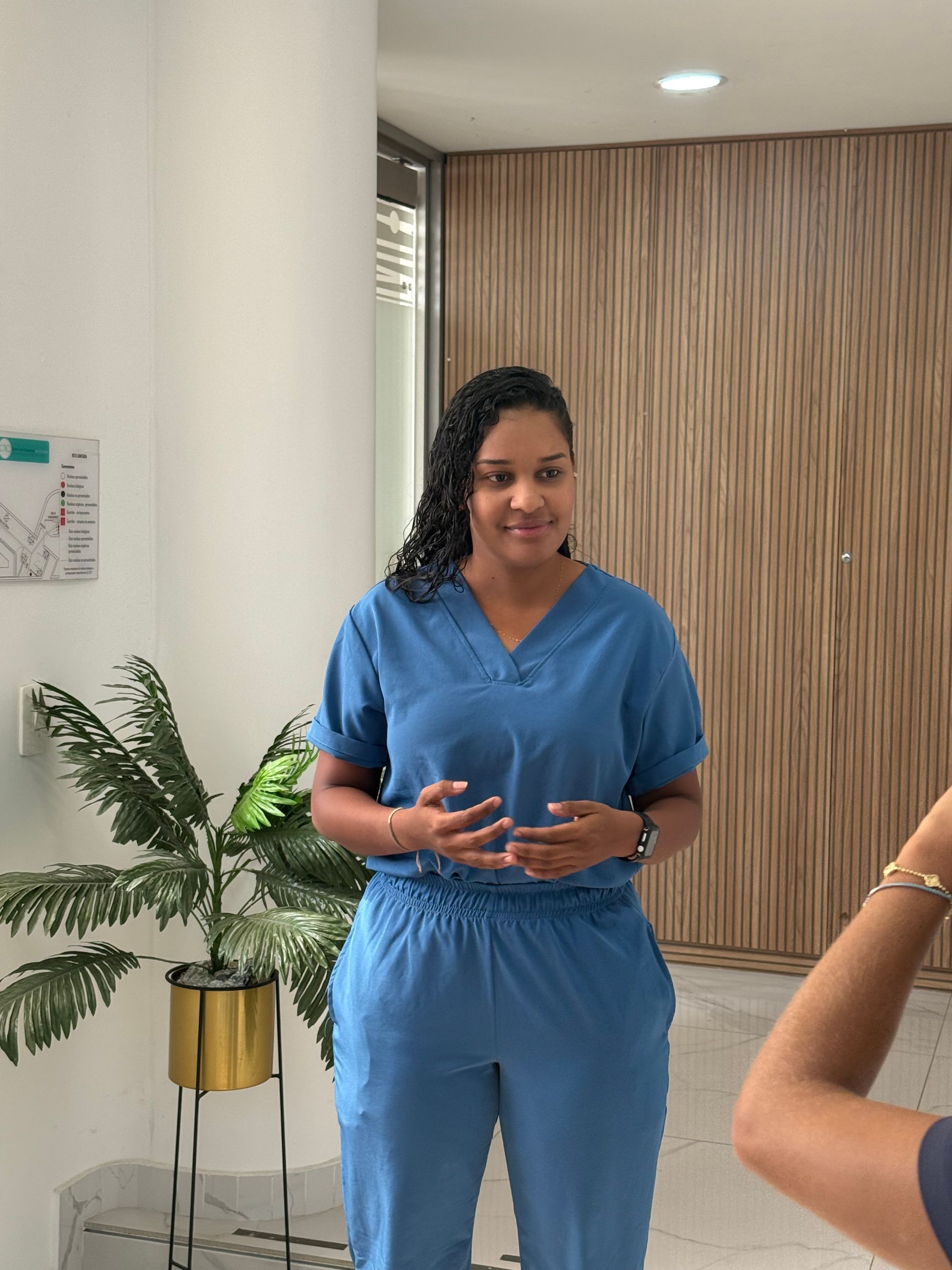 Woman in blue scrubs standing near a potted plant and a wooden wall, gesturing with hands.
