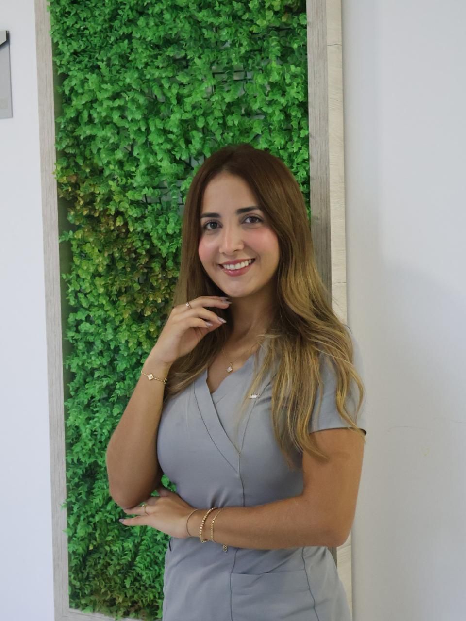 Woman in grey scrubs smiling, posing with hand near face, in front of a green wall and a white wall.