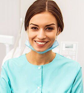 Dentist smiling, wearing mask around neck and teal scrubs, in an exam room.