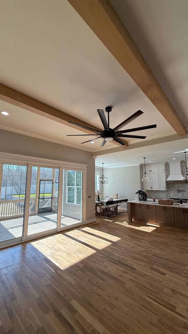 Spacious living area with wood floors, sliding glass doors, exposed beams, ceiling fan, and a kitchen in the background.