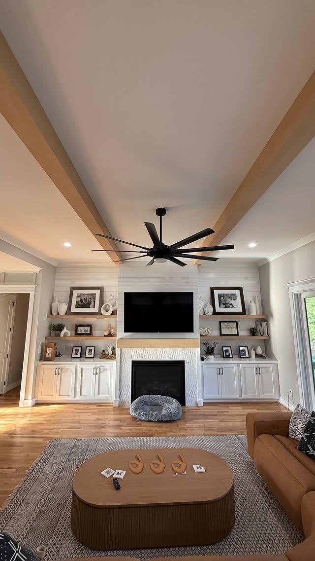 Living room with a fireplace, TV, and built-in shelves, adorned with a large ceiling fan and exposed beams.