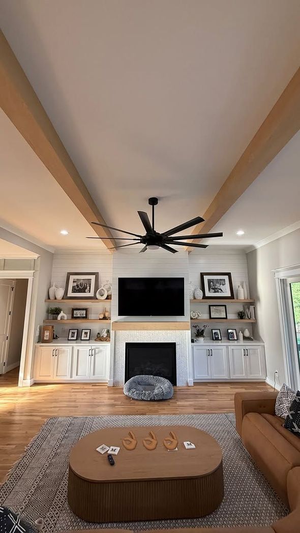 Living room with a fireplace, TV, and built-in shelves, adorned with a large ceiling fan and exposed beams.