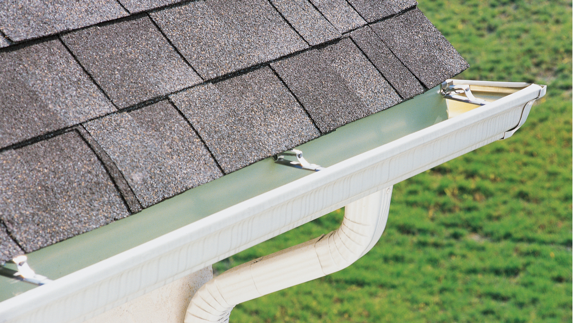 Gutter attached to roof with downspout, white with green roof, grass in background.