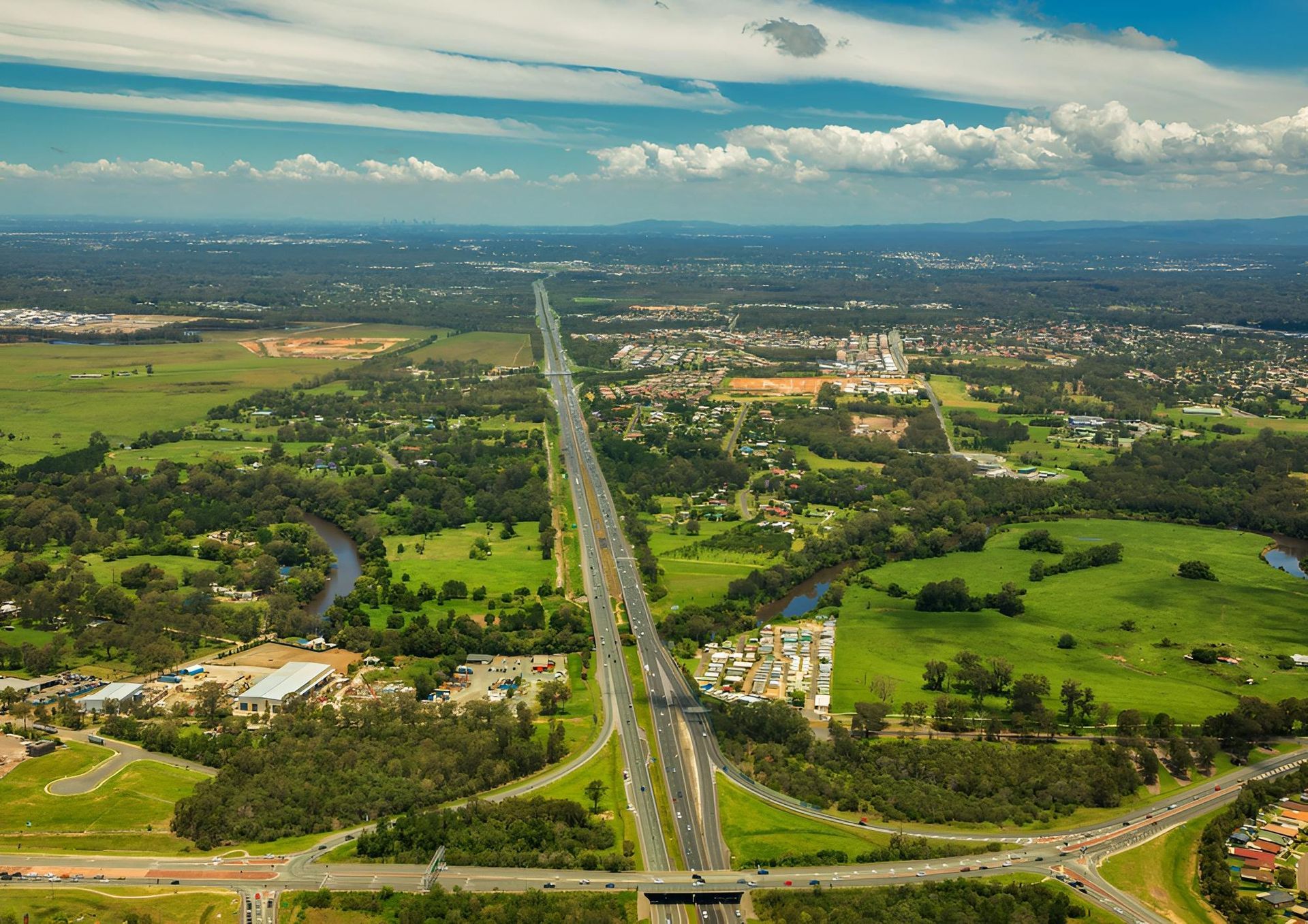 Aerial View of a Multi-lane Highway Cutting Through a Lush, Green Landscape With a River and Buildings in the Distance Under a Blue Sky — C & D Manufacturing In Caboolture, QLD