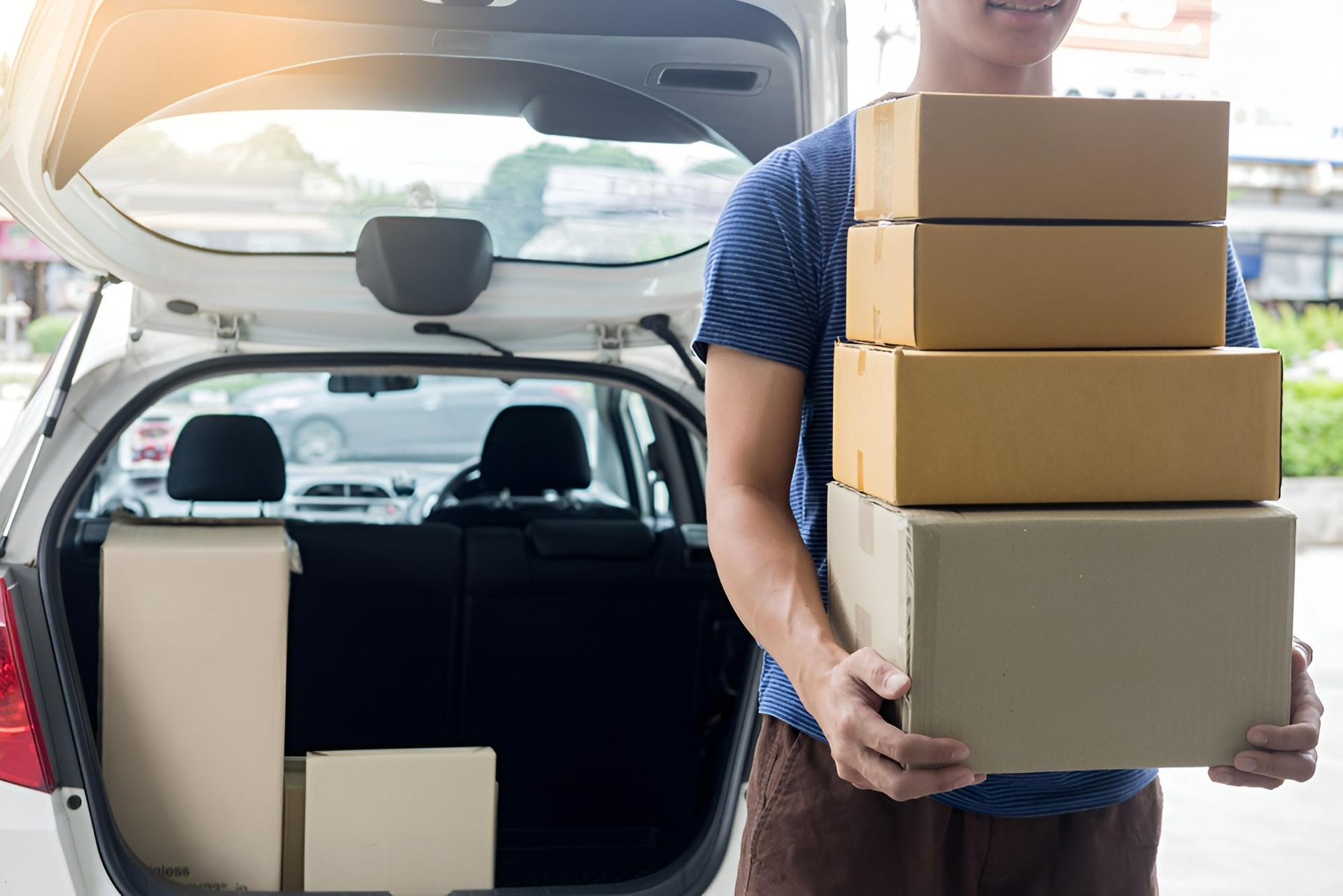 A Person Carrying a Stack of Cardboard Boxes From the Open Trunk of a White Car. They Are Smiling — C & D Manufacturing In Caboolture, QLD