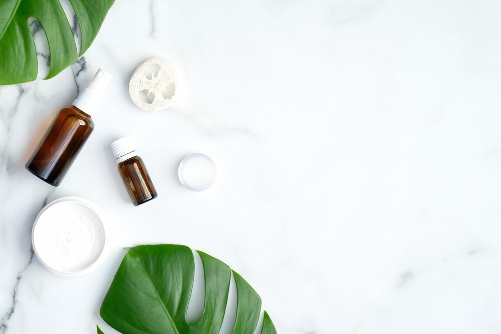 Brown Skincare Bottles, a White Powder Jar, and a Loofah Rest on a Marble Surface With Tropical Green Leaves — C & D Manufacturing In Caboolture, QLD