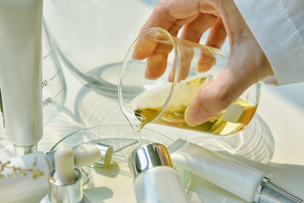 A Hand Pouring a Yellow Liquid From a Beaker Into Cosmetic Product Tubes on a White Surface — C & D Manufacturing In Caboolture, QLD