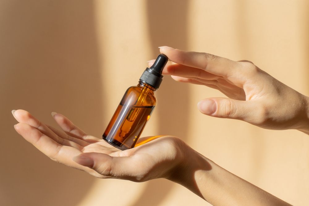 Hands Holding a Brown Glass Bottle With a Dropper, Against a Peach Background With Shadows — C & D Manufacturing In Caboolture, QLD