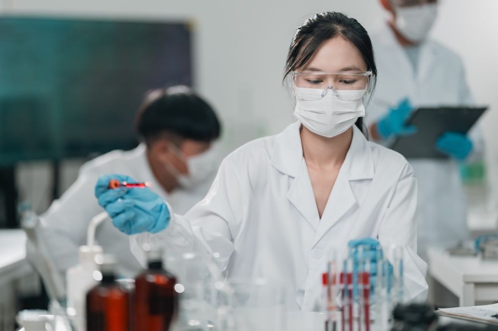 Laboratory Worker in a White Coat, Mask, and Goggles, Handling a Test Tube in a Lab. Other Researchers Are in the Background — C & D Manufacturing In Caboolture, QLD