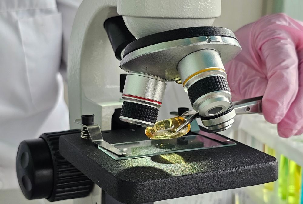 Person in a Lab Coat and Gloves Examines a Softgel Capsule With a Microscope in a Lab — C & D Manufacturing In Caboolture, QLD