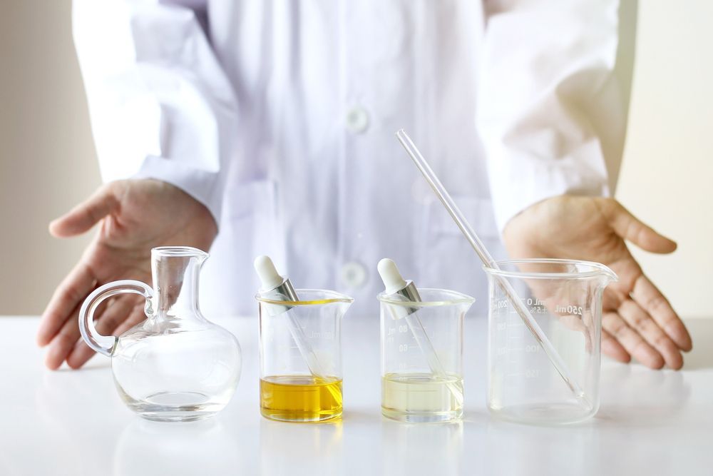 Person in Lab Coat Gestures Toward Beakers With Yellow and Clear Liquids and a Glass Flask on a White Surface — C & D Manufacturing In Caboolture, QLD