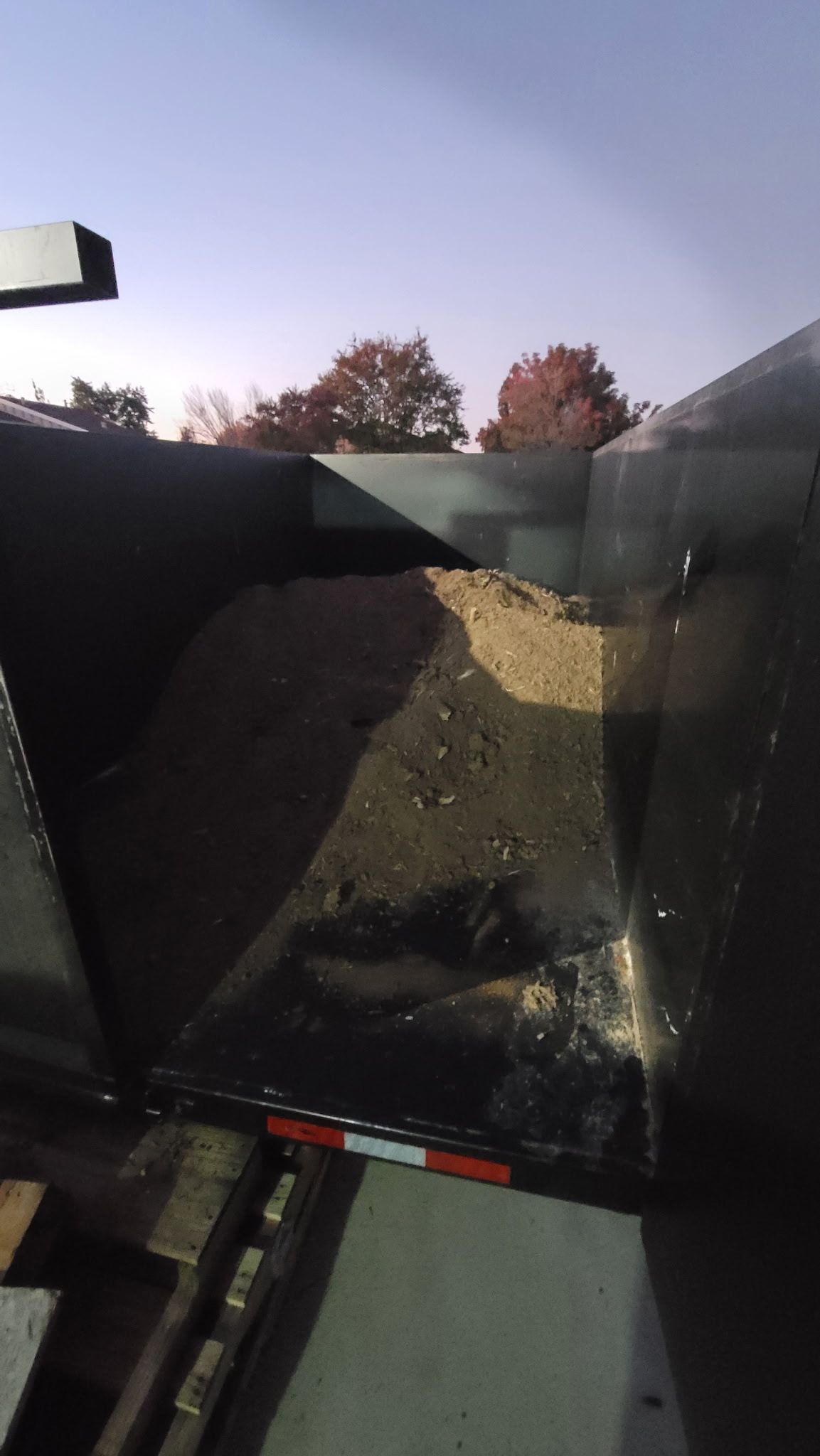 Dump truck bed filled with dirt and debris, with a bright sky and trees visible in the background.