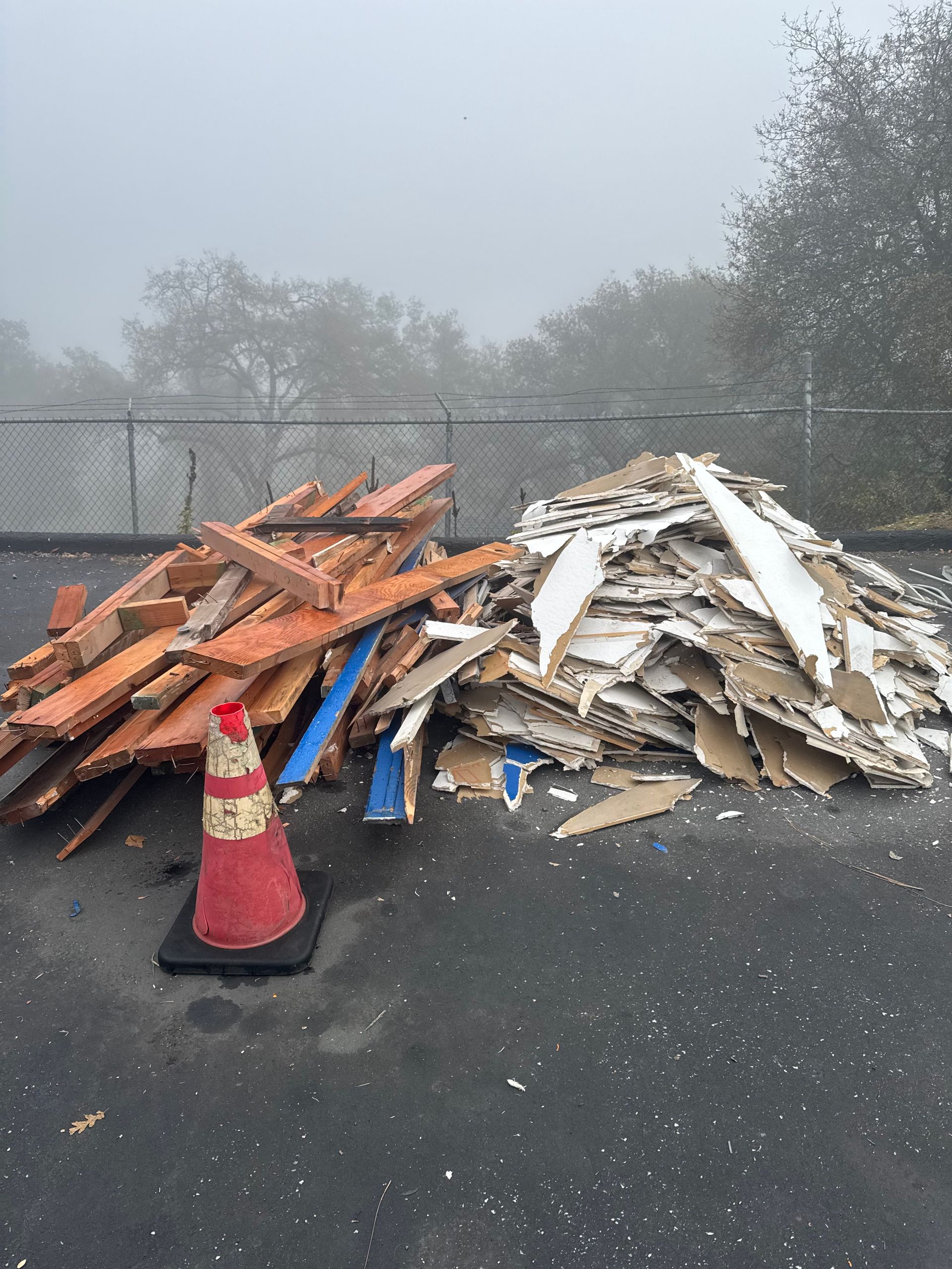 Piles of construction debris: orange and blue wood, white drywall, and a traffic cone, against a foggy backdrop.