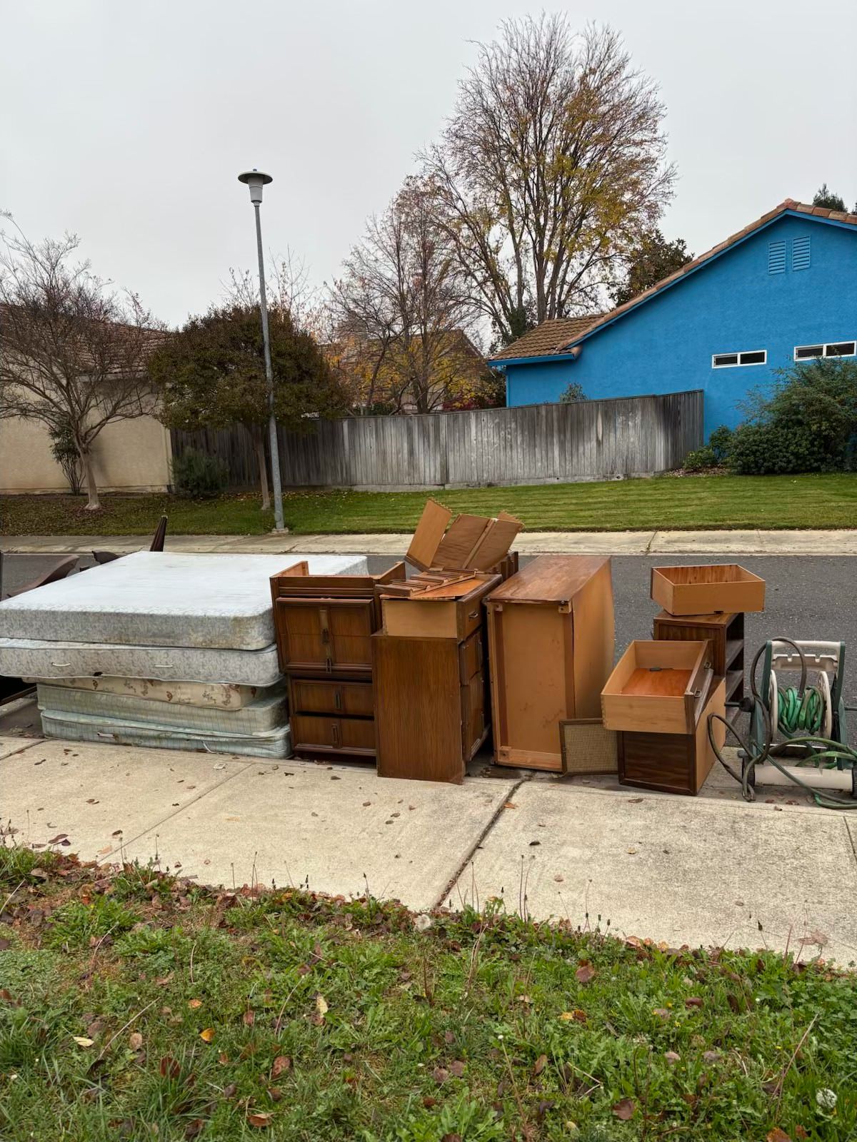 Pile of furniture and cardboard boxes on a sidewalk, next to a blue house and a street. Overcast sky.