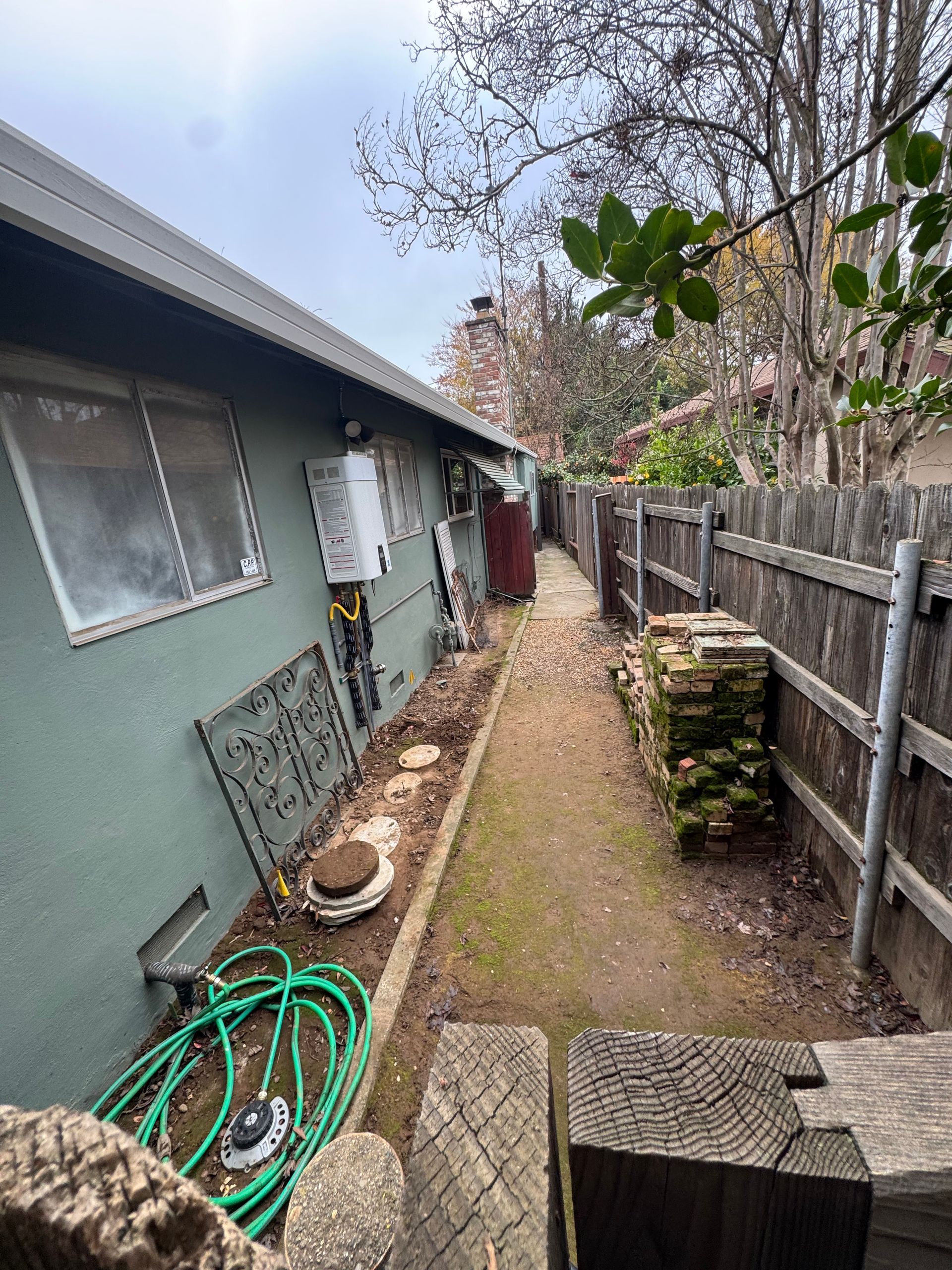Backyard pathway next to a green house with a fence on the right. Green hose and metal items are on the ground.