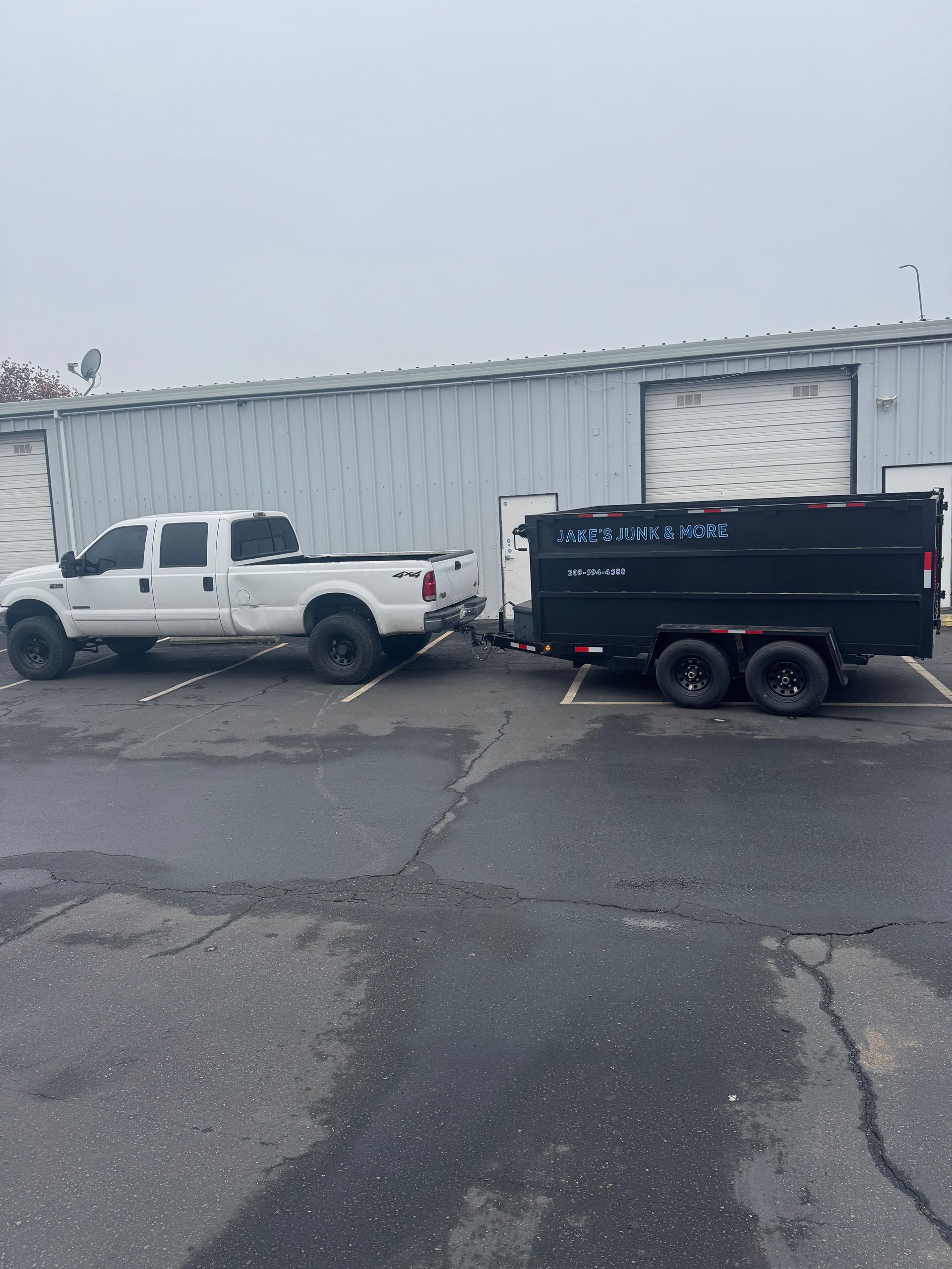 White pickup truck towing a black dump trailer in front of a gray building on a cloudy day.
