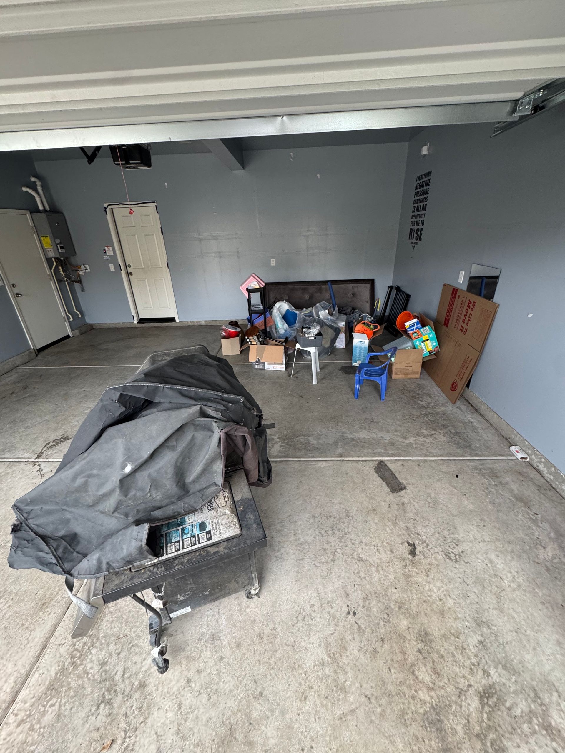 Cluttered garage with gray walls and concrete floor. Black tarp-covered object in foreground, boxes and items in center.
