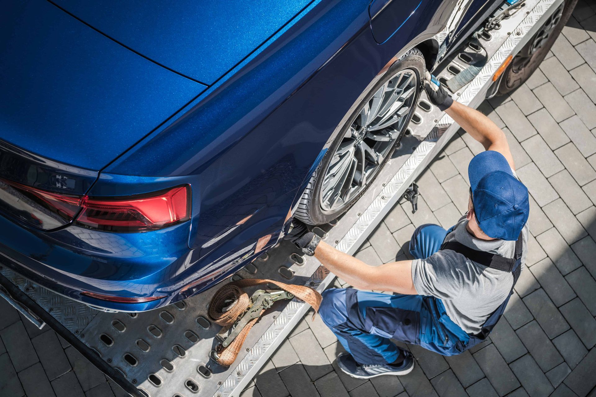 A person secures a blue car with straps on a car carrier in a paved lot.