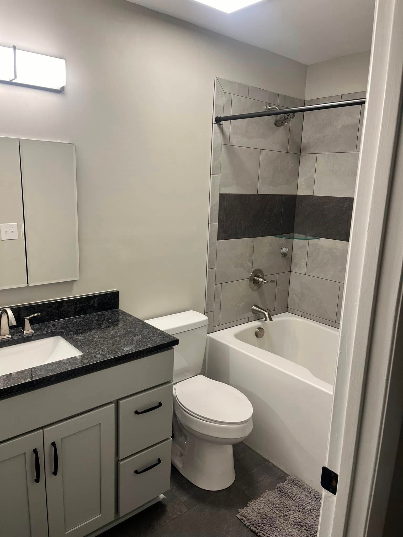 Bathroom with a gray vanity, white toilet, bathtub with gray tile, and a light gray wall.