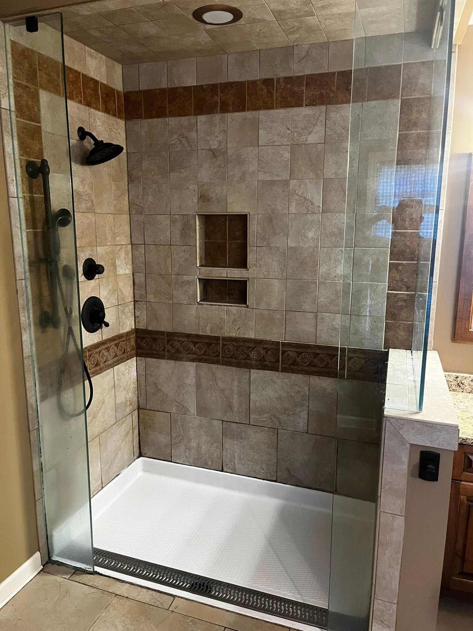 Tile shower with glass door and black fixtures. Beige and brown tiles with built-in shelf.