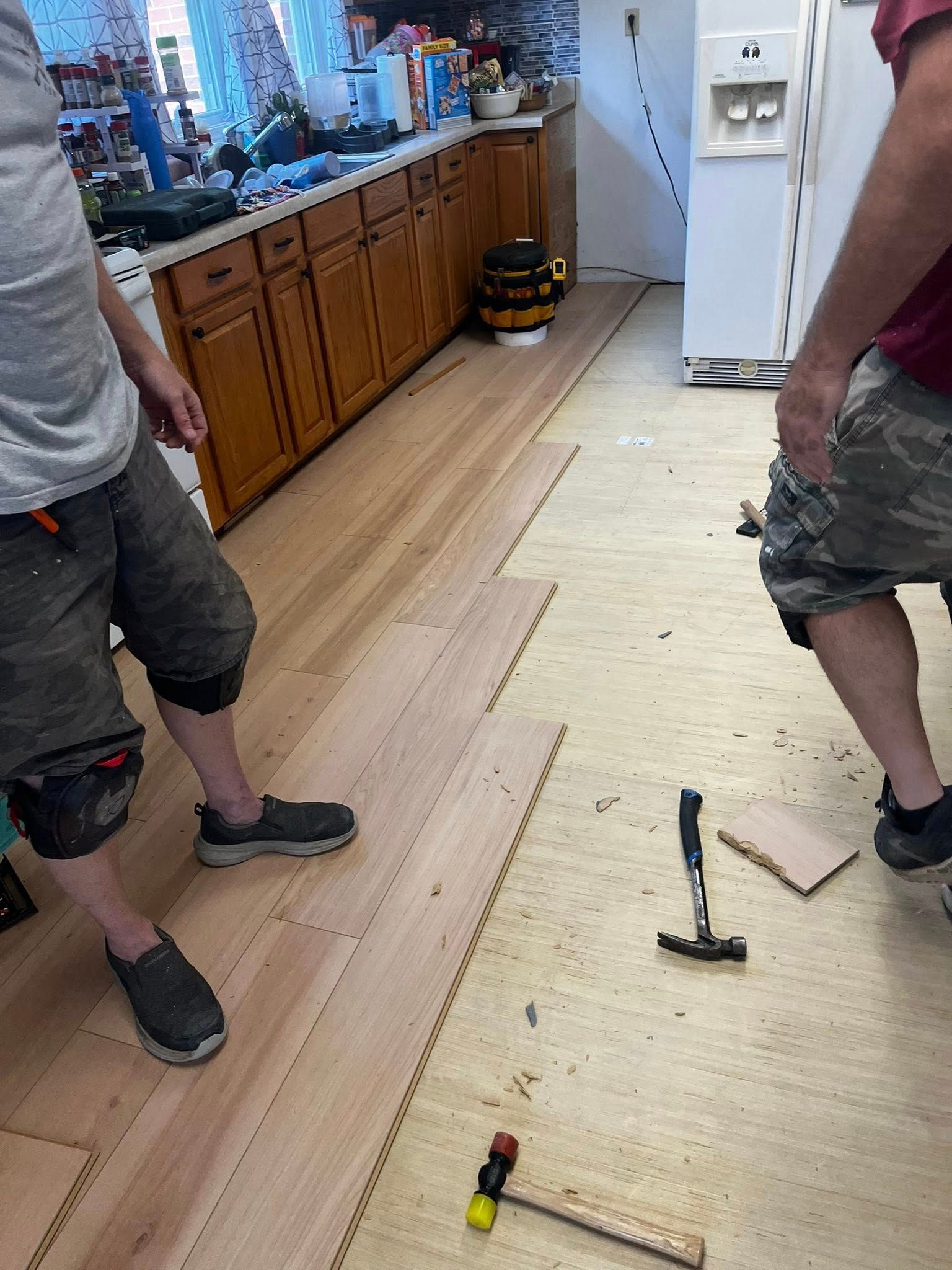 Two people installing hardwood flooring in a kitchen. Hammer and tools on the floor.